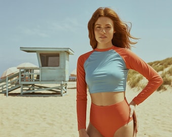 Op de afbeelding: Een tweedelig badpak met een lichtblauw top met lange mouwen en een hoog getailleerde oranje broek. De top heeft oranje mouwen en een lichtblauwe torso. Het badpak wordt gedragen op een zandstrand.