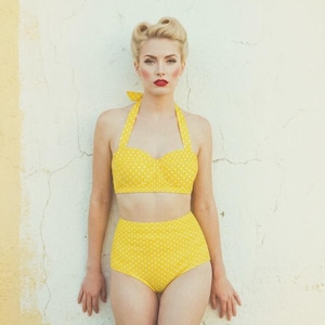 May include: A yellow polka dot bikini with a halter top and high-waisted bottoms. The two-piece swimsuit is set against a textured white and yellow wall. The model has blonde hair and red lipstick.
