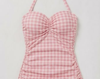 May include: A pink and white gingham-patterned swimsuit. The one-piece swimsuit features a halter neck tie, a sweetheart neckline, and ruching details. The bottom portion of the swimsuit is a solid light pink color.