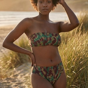 May include: A tie-dye bikini set in green and burgundy. The strapless top and high-waisted bottoms are made of a soft, stretchy fabric. The model is standing on a sandy beach with tall grass in the background. The bikini is a summer swimwear item.