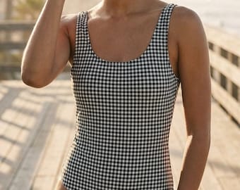 May include: A one-piece swimsuit with a black and white gingham pattern. The scoop-neck swimsuit is worn by a person wearing black cat-eye sunglasses. The person is standing on a wooden boardwalk near the beach.