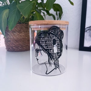 Könnte beinhalten: Ein klares Glasgefäß mit einem Holzdeckel, das eine schwarz-weiße Illustration einer Frau mit einem Bücherregal als Kopf zeigt. Die Frau greift nach einem Buch auf einer Leiter.