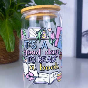 Könnte beinhalten: Ein Glasgefäß mit Bambusdeckel, das mit einer bunten Illustration eines Buches, Blumen und dem Text "Es ist ein guter Tag, um ein Buch zu lesen" verziert ist.