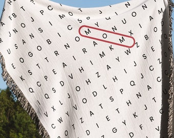 May include: A white woven blanket with a black and white word search pattern. The word "MOM" is highlighted in red. The blanket is being held up by a person in a grassy field.