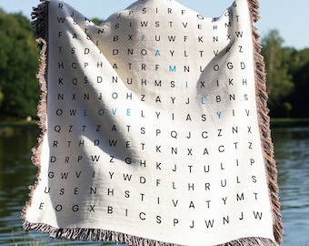 May include: A white throw blanket with a woven design featuring the word "LOVE" and the word "FAMILY" in blue. The blanket is being held by a person standing in a grassy area near a body of water.