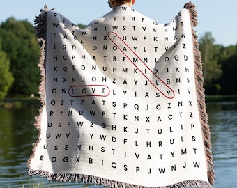 May include: A white throw blanket with a black and red word search pattern. The word "LOVE" is highlighted in red. The blanket is being held by a person standing in a grassy area near a body of water. The text "Also available as Minky Blanket" is visible at the bottom of the image.