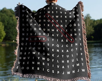 May include: A black woven throw blanket with a white word search pattern that spells out the word "LOVE" in red. The blanket is draped over a person's shoulders.