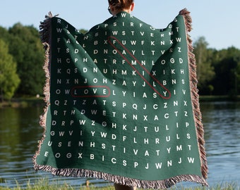 May include: A green throw blanket with a woven word search pattern. The word "LOVE" is highlighted in red. The blanket is being held by a person standing in front of a body of water.