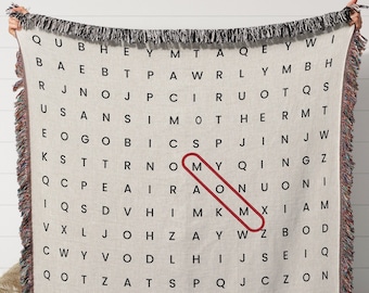 May include: A woven throw blanket with a beige background and a black and white word search puzzle. The word "MOM" is highlighted in red.
