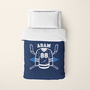 May include: Navy blue twin-sized duvet cover featuring a white hockey jersey design. The jersey has the name "ADAM" and the number "88" on it. Two white hockey sticks are crossed behind the jersey.