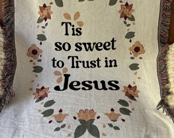 May include: A white woven throw blanket with a floral border and the text "Tis so sweet to Trust in Jesus".