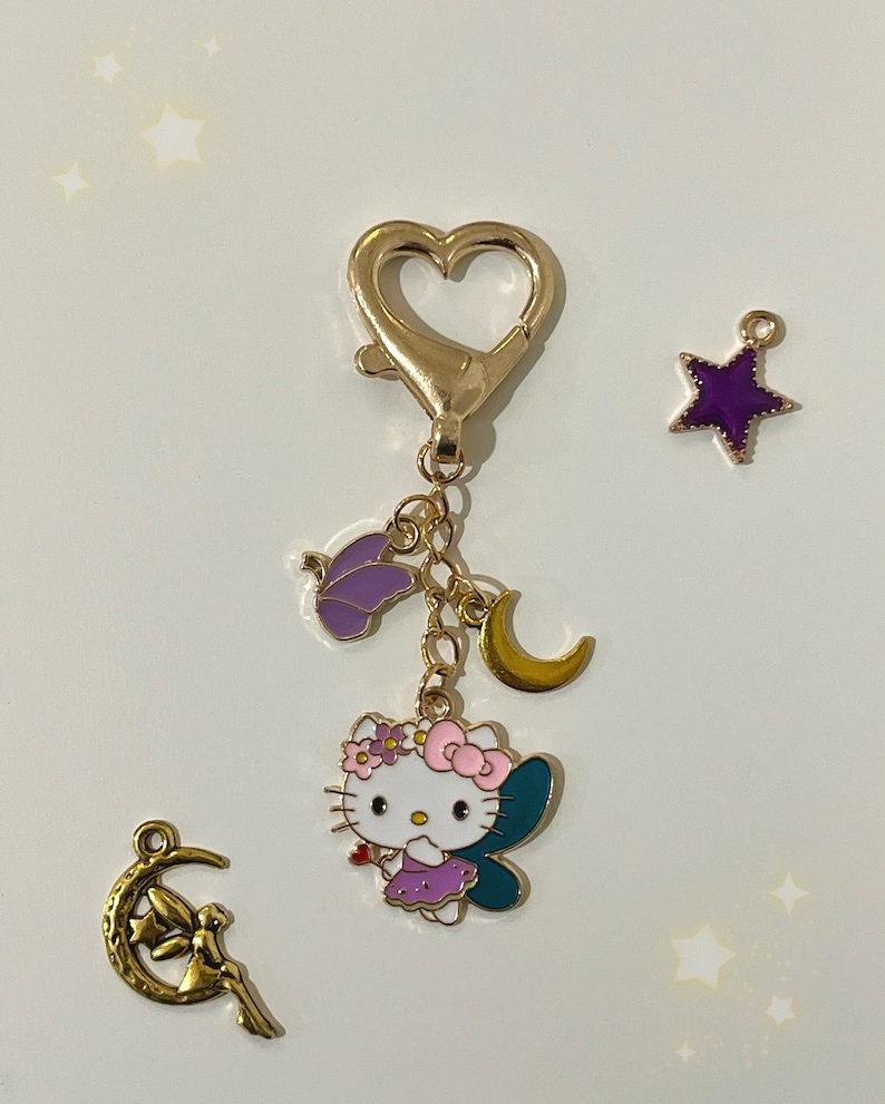 May include: A gold-tone keychain featuring a Hello Kitty fairy charm with purple wings, a flower crown, and a pink bow. The keychain also includes a purple star charm, a gold crescent moon charm, and a butterfly charm.