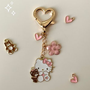 Peut inclure: Porte-clés en or avec un charme Hello Kitty rose tenant un charme ours en peluche brun. Le porte-clés a également des charmes de fleurs et de cœurs roses.