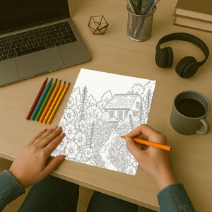 May include: A person coloring a detailed line drawing of a house and garden with an orange pencil. A laptop, headphones, and a mug of coffee are on the desk. Colored pencils are arranged in a row.