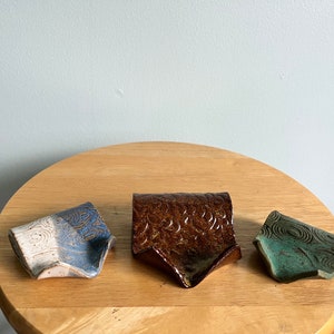 May include: Three ceramic soap dishes with different patterns and colors. The dish on the left is white with blue swirls, the middle dish is brown with a floral pattern, and the dish on the right is green with a swirl pattern.