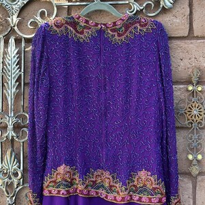 May include: A long-sleeved, purple dress with intricate beadwork. The dress features a detailed pattern of silver beads across the bodice and sleeves. The neckline and hem are adorned with colorful, ornate embroidery in shades of pink, gold, and green.