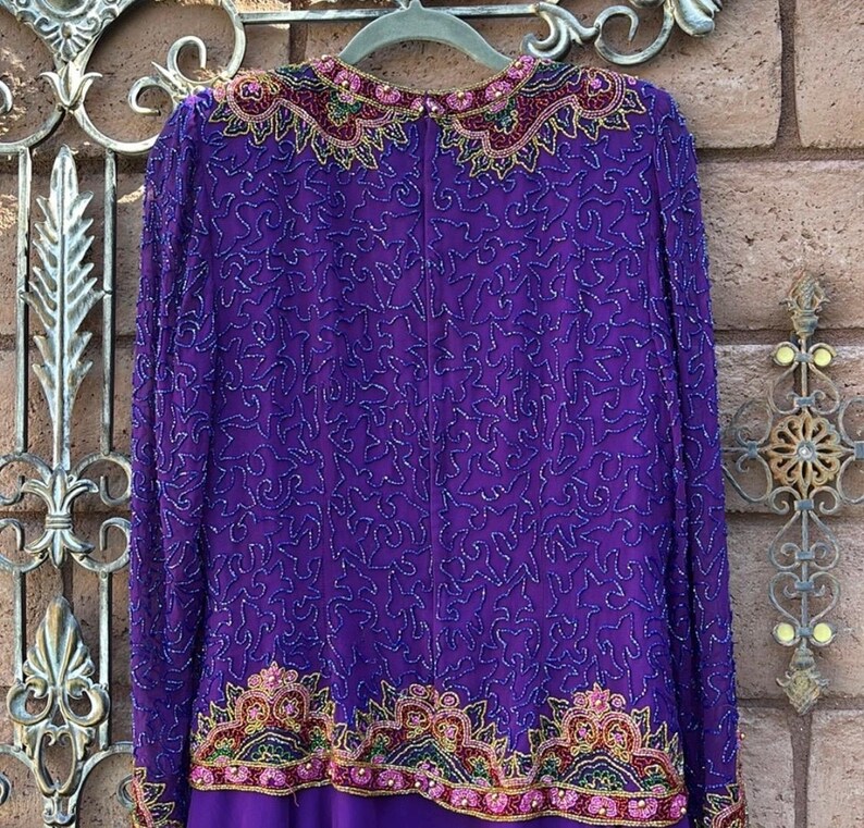 May include: A long-sleeved, purple dress with intricate beadwork. The dress features a detailed pattern of silver beads across the bodice and sleeves. The neckline and hem are adorned with colorful, ornate embroidery in shades of pink, gold, and green.
