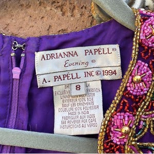 May include: Close-up of a purple Adrianna Papell evening dress tag. The tag reads "Adrianna Papell Evening" and "A. Papell Inc &copy; 1994". The dress has a beaded neckline with pink and gold accents. The zipper is purple.