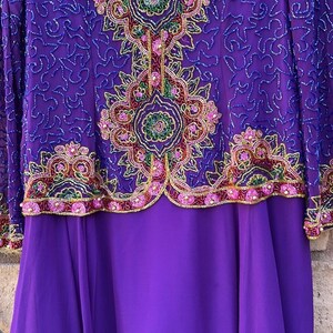 May include: A long purple dress with intricate beadwork and embroidery. The bodice features a detailed floral design in shades of green, pink, and gold. The sleeves are sheer, and the skirt is a solid, flowing purple fabric. The dress is a formal garment.