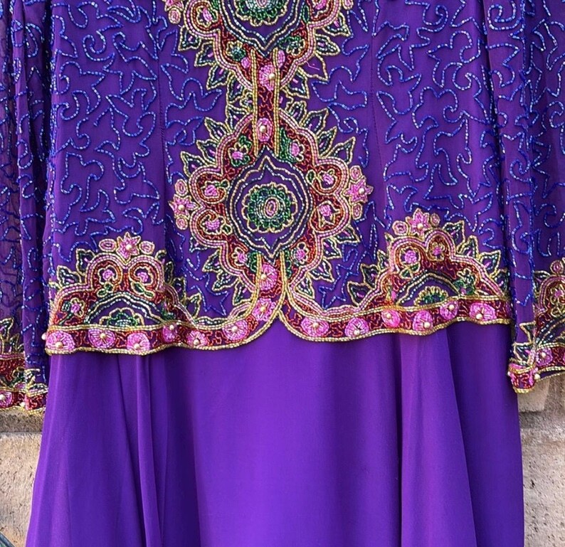 May include: A long purple dress with intricate beadwork and embroidery. The bodice features a detailed floral design in shades of green, pink, and gold. The sleeves are sheer, and the skirt is a solid, flowing purple fabric. The dress is a formal garment.