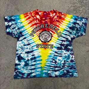 May include: A tie-dye t-shirt with a vibrant mix of colors including red, yellow, blue, and purple. The shirt features the text "PSYCHEDELIC STATE" in a circular design with additional text and a graphic.