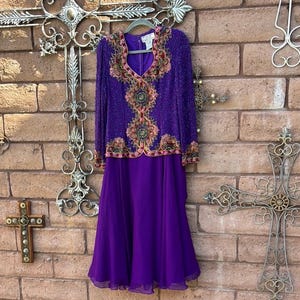 May include: A two-piece purple formal dress. The long-sleeved top is heavily embellished with beads and embroidery in shades of pink, gold, and green. The skirt is a flowing, solid purple. The dress hangs on a hanger against a brick wall.
