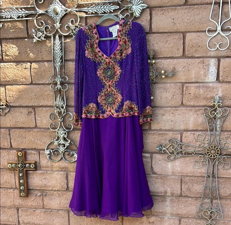 May include: A two-piece purple formal dress. The long-sleeved top is heavily embellished with beads and embroidery in shades of pink, gold, and green. The skirt is a flowing, solid purple. The dress hangs on a hanger against a brick wall.