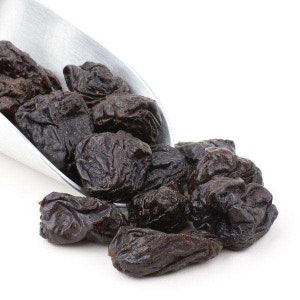May include: A metal scoop filled with dried prunes, a type of dried plum, spilling out onto a white surface.