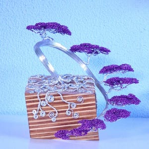 Op de afbeelding: Handgemaakte draadsculptuur van een bonsai-boom. De boom heeft een zilveren draadstam en takken met levendig paars draadbladeren. De boom staat op een houten blok met een gestreept patroon, wat een uniek decoratief stuk creëert.