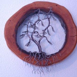 Op de afbeelding: Handgemaakte draadboomsculptuur met een gedetailleerde stam, takken en wortels. De boom is geplaatst in een ronde, natuurlijke houten lijst. De houten lijst heeft een rijke bruine kleur en een zichtbare houtnerf.