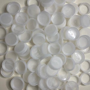 Puede incluir: Un conjunto de tapones de botella de plástico blanco, algunos con bordes texturizados, dispersos sobre una superficie. Los tapones son redondos con una parte superior plana y un borde ligeramente elevado. La imagen muestra una variedad de tapones.