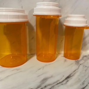 以下が含まれることがあります： 白いスクリューキャップが付いた、オレンジ色の処方薬ボトルが3つ。ボトルはサイズが異なり、左が最大、右が最小です。ボトルは空で透明です。