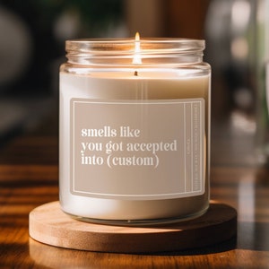 May include: A white candle with a clear glass jar and a label that reads "smells like you got accepted into (custom)". The candle is lit and sitting on a wooden coaster.
