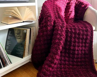 May include: A plush, burgundy-colored blanket draped over a white wicker chair. The blanket has a textured, raised pattern. A white bookshelf with a stereo, open book, and vinyl records is in the background.