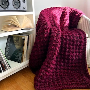 Puede incluir: Una manta de color burdeos con un patrón texturizado, sobre una silla blanca. En el fondo, una estantería blanca con un equipo de música, un libro abierto y discos de vinilo.