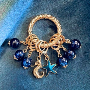 Può includere: Un anello in metallo dorato con una trama intrecciata, adornato con ciondoli pendenti. I ciondoli includono perle di vetro blu scuro e ciondoli a tema marino: un cavalluccio marino e una stella marina. Lo sfondo è un tessuto verde acqua strutturato.