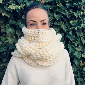 以下が含まれることがあります： 人の首に巻かれた白いクロシェの無限スカーフ。スカーフは厚くてずっしりしていて、その人は白いセーターを着ています。