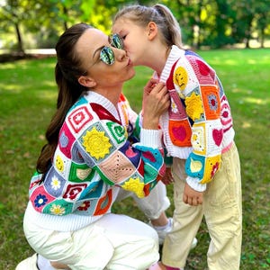 May include: A woman and a child are seen wearing vibrant, patchwork crochet cardigans. The cardigans display squares with diverse motifs, such as flowers, hearts, and smiley faces. The woman is wearing white trousers and sunglasses, the child is in khaki trousers.