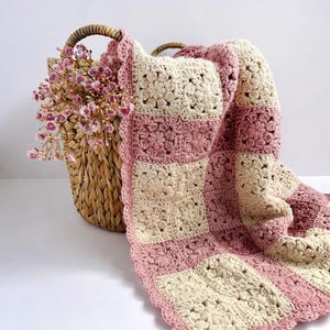 May include: A handmade crochet blanket in a floral pattern, featuring alternating squares of cream and light pink. The blanket is draped over a woven basket with a handle, which contains pink floral accents. The blanket's edge has a scalloped trim.