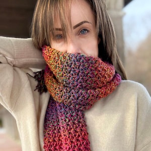 May include: A colorful crocheted scarf with a gradient of red, orange, yellow, green, blue, and purple. The scarf is worn around the neck of a person.