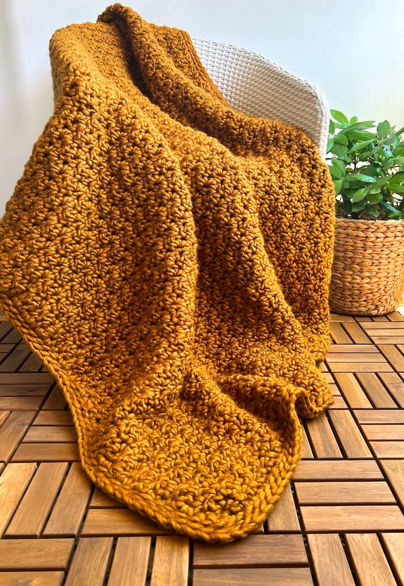 May include: Mustard yellow crocheted blanket draped over a white chair. The blanket has a textured stitch pattern and is made of thick yarn. A potted plant in a woven basket sits nearby on a wooden deck.