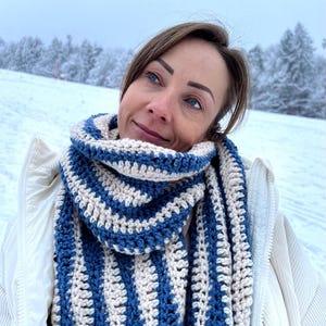 Puede incluir: Una mujer con una chaqueta blanca y una bufanda de ganchillo a rayas azules y blancas. La bufanda es larga y tiene un diseño sencillo.
