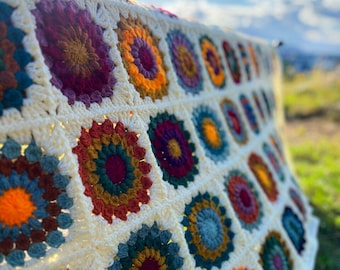 May include: A handmade crochet blanket featuring a repeating pattern of circular floral motifs in vibrant shades of orange, yellow, deep red, purple, and teal, all joined together by a cream-colored yarn border in a classic granny square style.