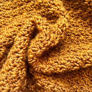 May include: Close-up of a mustard-colored, chunky knit textile. The fabric is densely woven with a textured, looped pattern, creating a soft, warm appearance. The folds and shadows add depth and dimension to the knitted material, suggesting a cozy blanket or scarf.
