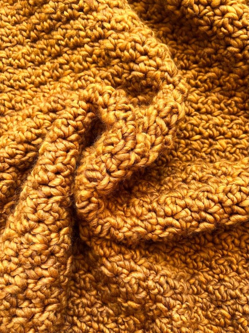May include: Close-up of a mustard-colored, chunky knit textile. The fabric is densely woven with a textured, looped pattern, creating a soft, warm appearance. The folds and shadows add depth and dimension to the knitted material, suggesting a cozy blanket or scarf.