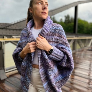 Può includere: Un cardigan in crochet a righe viola e blu con collo a lupetto e maniche lunghe. Il cardigan è indossato sopra una camicia bianca e pantaloni bianchi.