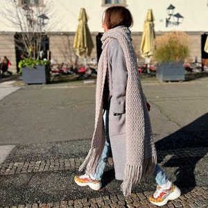 May include: A woman wearing a long, light gray knitted scarf with fringe, a gray coat, and blue jeans. She is wearing colorful sneakers.