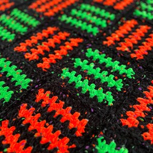 May include: Close-up of a handmade crochet blanket with a repeating pattern. The blanket features a black background with vibrant neon green and orange zig-zag designs. The yarn has a textured appearance.