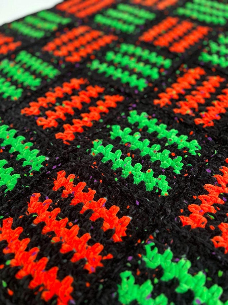 May include: Close-up of a handmade crochet blanket with a repeating pattern. The blanket features a black background with vibrant neon green and orange zig-zag designs. The yarn has a textured appearance.