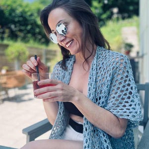 Op de afbeelding: Een lichtblauwe, open geweven gehaakte sjaal. De sjaal wordt gedragen over een zwarte top en witte shorts. De persoon draagt een zonnebril en houdt een glas rode drank met een zwart rietje vast. De achtergrond omvat groene bomen en een blauwe lucht.